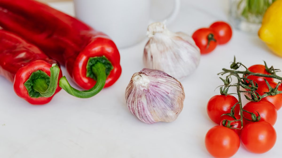 Fresh garlic placed among ripe red cherry tomatoes and peppers on white table near white ceramic cup and yellow lemon